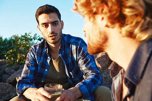Waist-up portrait of handsome bearded man with glass of cocktail in hands talking to his red-haired friend while enjoying summer day outdoors