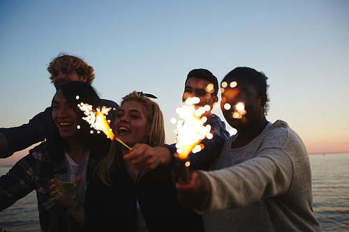 Male and female friends with sparklers in hands gathered together outdoors and celebrating momentous event, picturesque seascape on background