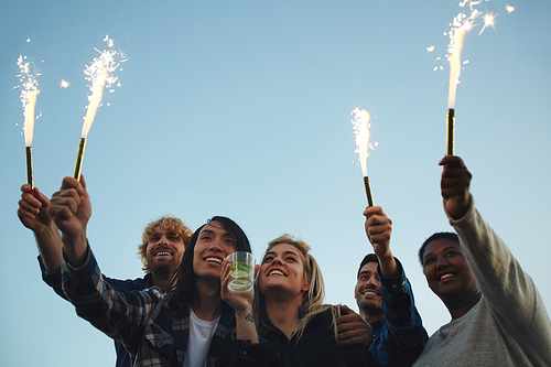 Low angle view of multi-ethnic group of cheerful friends looking into distance with toothy smiles while lighting sparklers at outdoor party