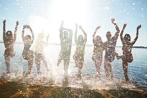 Playful teens raising hands while splashing water by sand