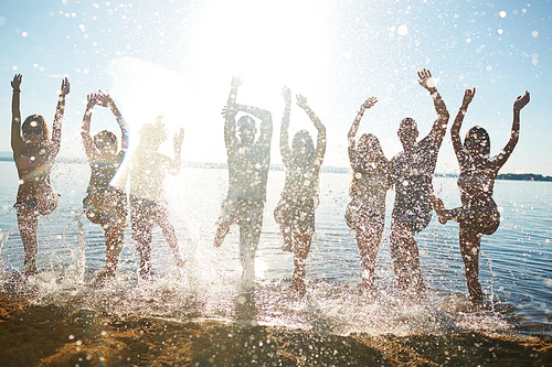Playful teens raising hands while splashing water by sand