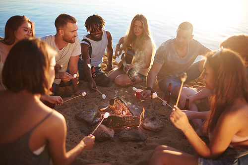 Group of restful friends roasting marshmallows while sitting by bonfire