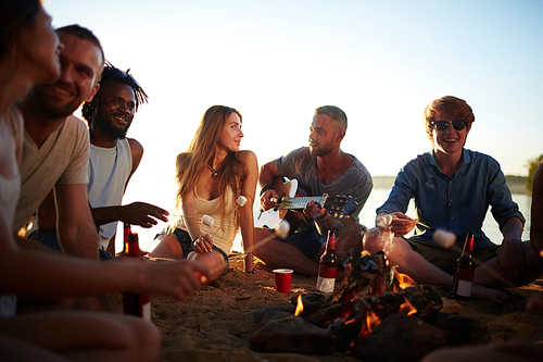 Friendly guys and girls spending summer evening around campfire