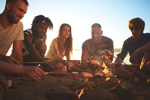 Young buddies roasting sausages on campfire by seaside