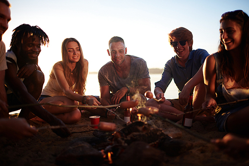 Friendly girls and guys roasting sausages on campfire on the beach