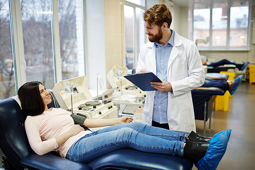 Girl sitting in arm-chair and talking to doctor while donating blood