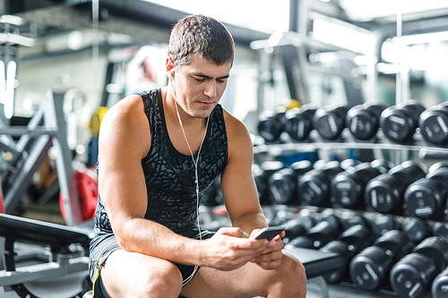 Portrait of strong muscular man  listening to music using smartphone while sitting on bench before work out  in gym