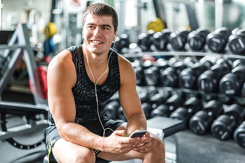 Portrait of strong muscular man smiling and  listening to music using smartphone while sitting on bench before work out  in gym