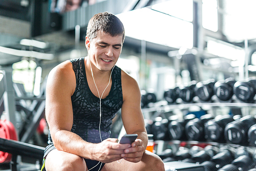 Portrait of strong muscular man  listening to music looking at  smartphone screen while sitting on bench before work out  in gym