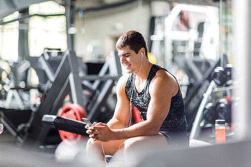 Portrait of strong muscular man  listening to music looking at  smartphone screen while sitting on bench in machines hall of gym
