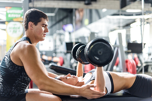 Portrait of muscular personal instructor helping beautiful woman doing weightlifting exercise with big dumbbells on bench  in modern gym