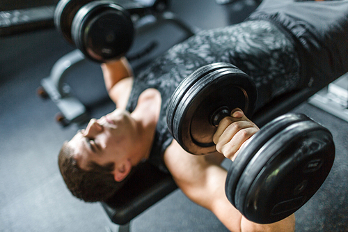 High angle view portrait of muscular man doing bench press exercise for chest muscles using heavy dumbbells in modern gym