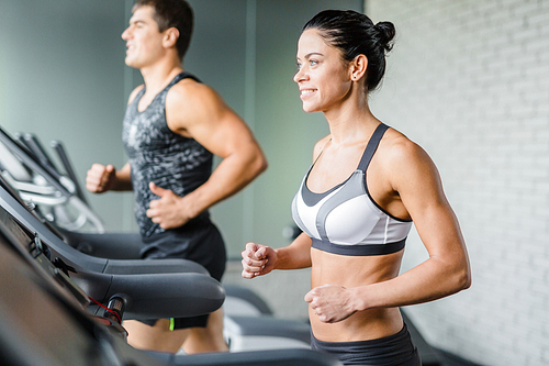 Portrait of beautiful   sportive brunette woman smiling while exercising on treadmill in gym next to fit man