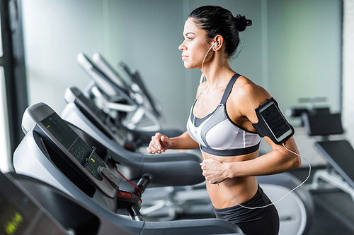 Portrait of sportive brunette woman exercising on treadmill in gym listening to music using shoulder smartphone holder