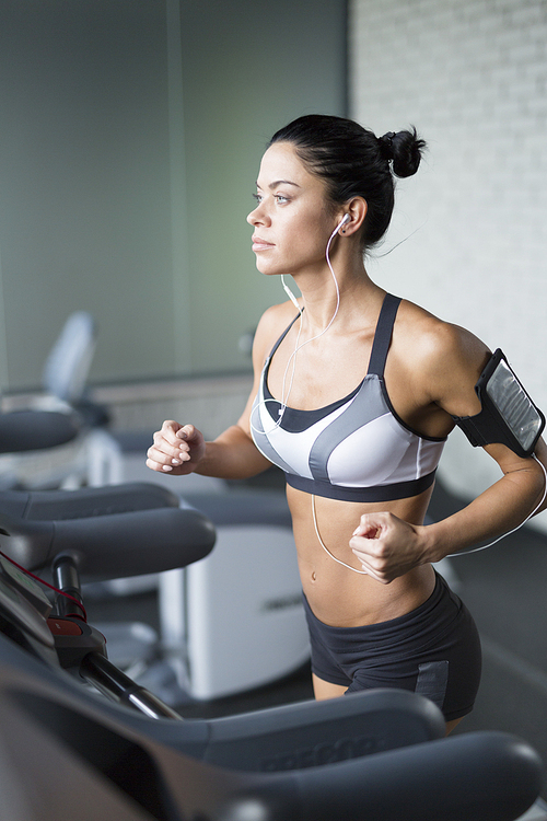 Portrait of sportive brunette woman  exercising on treadmill in gym listening to music using shoulder smartphone holder