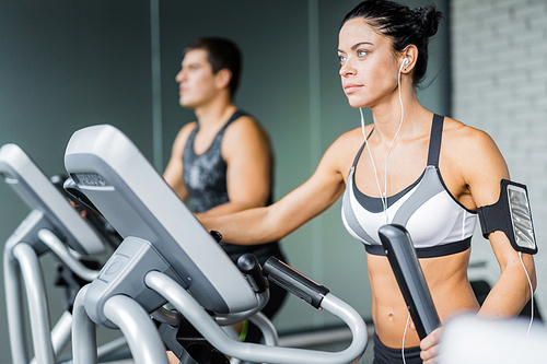 Portrait of beautiful  sportive brunette woman exercising using elliptical machine and listening to music next to fit man during workout in modern gym