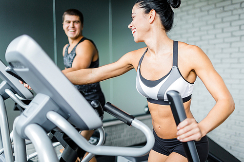 Portrait of beautiful  sportive brunette woman exercising using elliptical machine next to fit man and smiling to him during workout in modern gym