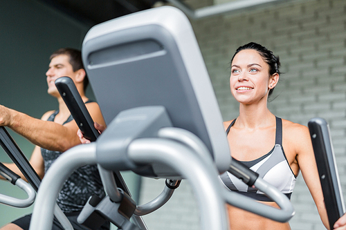 Low angle portrait of beautiful  sportive brunette woman  exercising using elliptical machine  next to fit man, both smiling during workout in modern gym