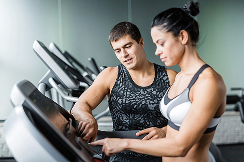 Portrait of muscular personal instructor explaining to fit woman how to use elliptical machine for exercise in modern gym