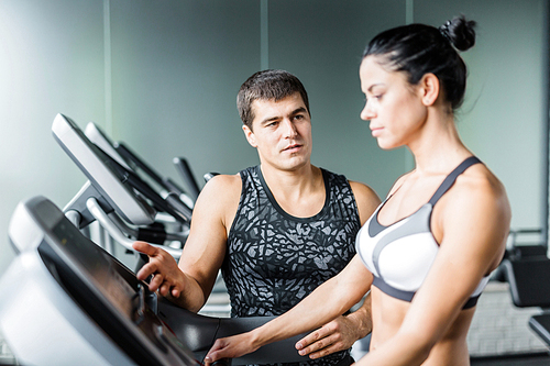 Portrait of muscular personal instructor explaining to fit woman how to set up elliptical machine for exercise in modern gym