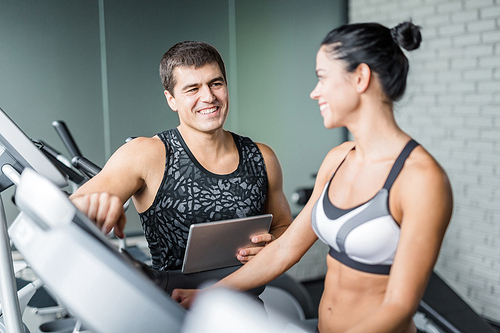 Portrait of muscular personal instructor watching smiling fit woman use elliptical machine for exercise in modern gym