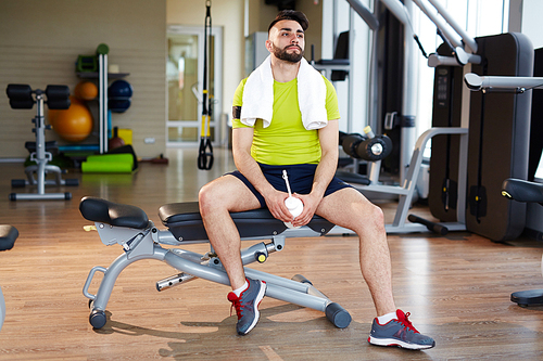 Working out in gym: handsome muscular man, wearing bright green sportsclothes,  sitting on adjustable bench resting after practice with machines