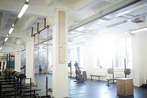 Background image of various equipment in gym: exercise stands with climbing rope and barbell benches in empty sunlit hall
