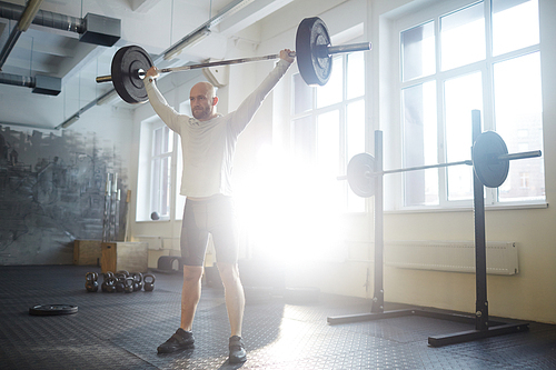 Wide shot portrait of modern bald strongman lifting heavy barbell overhead during workout in sunlit gym