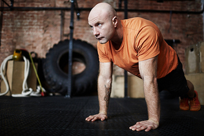 Handsome middle-aged sporty man wrapped up in intensive training: he flexing muscles while doing push-ups in gym, full-length portrait