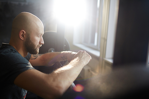 Side view portrait of modern bald bearded man looking out of window pensively in gym, leaning on weights rack in sunlight