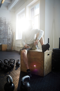 Portrait of modern sportsman sitting on wooden box in sunlit gym using smartphone