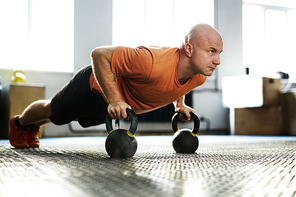 Intensive training in modern gym illuminated with bright sunlight: bald middle-aged sportsman standing in plank position with help of kettlebells, full-length portrait