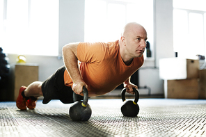 Concentrated fit man doing push-ups with kettlebells against white wall with panoramic windows of modern gym, full-length portrait