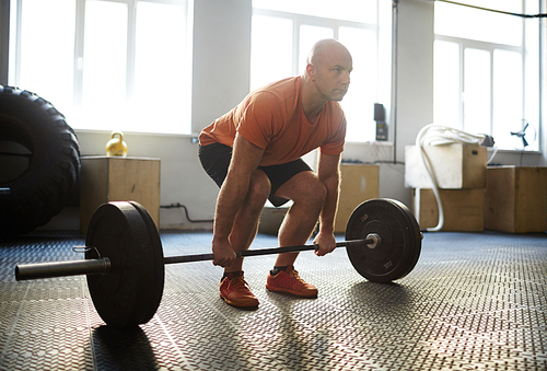 Bald middle-aged sportsman doing squats with barbell in spacious gym, full-length portrait