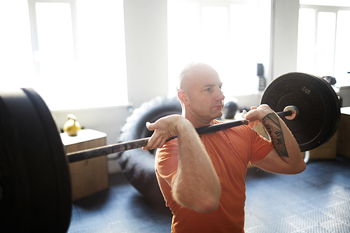 Concentrated bodybuilder doing shoulder press exercise with barbell in gym illuminated with bright sunlight