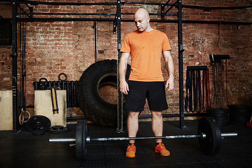 Full-length portrait of handsome fit man looking at barbell with concentration while standing in modern gym