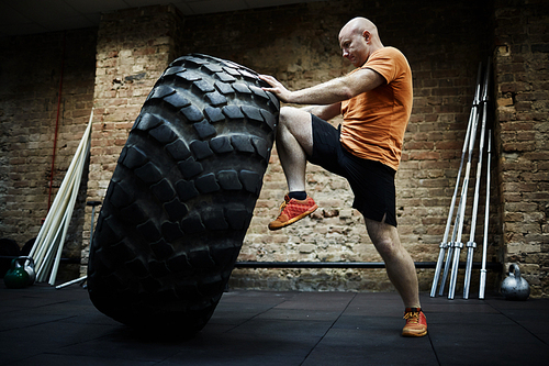 Confident bald sportsman exercising with huge tire in spacious dim gym, profile portrait shot