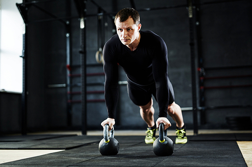 Intense crossfit workout in gym: male athlete performing kettlebell pushups looking determined and strained