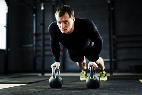 Powerful adult man wearing black sportswear performing kettlebell pushups in modern crossfit gym
