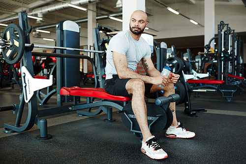 Portrait of handsome muscular sportsman sitting on weights bench in modern gym, looking away