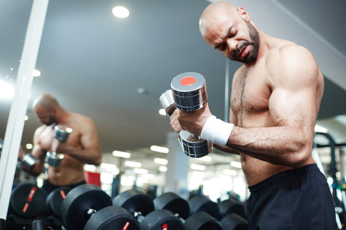 Portrait of shirtless muscular man doing arm exercise working out with dumbbells during strength training in modern gym, flexing and pumping bicep muscles