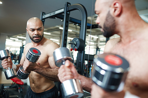 Portrait of shirtless muscular man looking in mirror, doing arm exercise working out with dumbbells during strength training in modern gym
