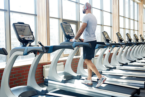 Back view portrait of muscular sportsman running on treadmill listening to music  alone in empty sunlit gym