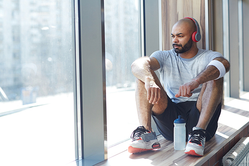 Portrait of contemporary muscular sportsman sitting by window listening to music in big headphones and looking away  pensively, resting after gym workout in sunlight
