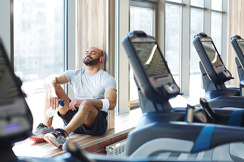 Portrait of modern trendy  sportsman sitting  in sunlight by window listening to music in big headphones with eyes closed, resting after gym workout behind row of treadmills