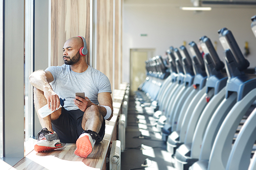 Calm man in activewear listening to music in headphones while looking in window of gym