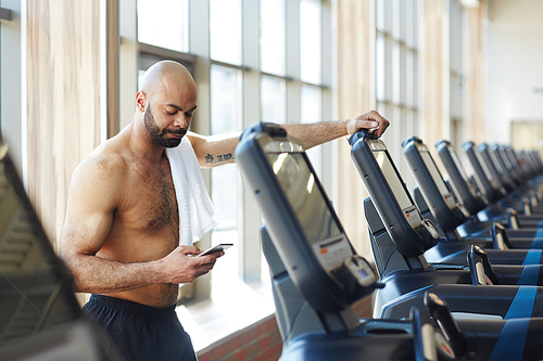 Young sporty man leaning on treadmill and texting in smartphone