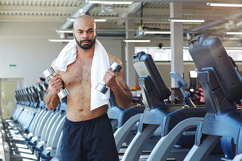 Shirtles man with towesl and dumbbells exercising on background of treadmills