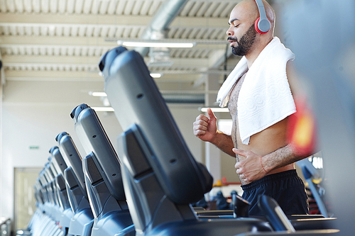 Side view portrait of muscular sportsman running on treadmill listening to music during intense cardio workout in modern gym