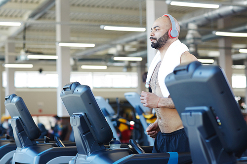 Side view portrait of muscular sportsman running on treadmill listening to music during cardio workout in modern gym